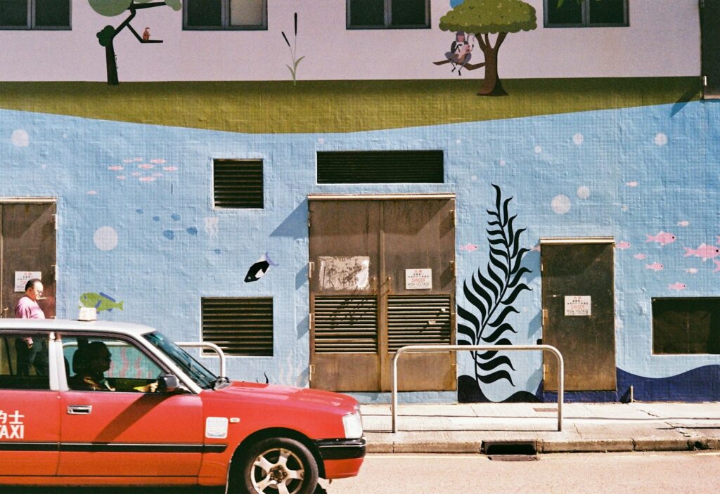 Red taxi passes a building with a mural.